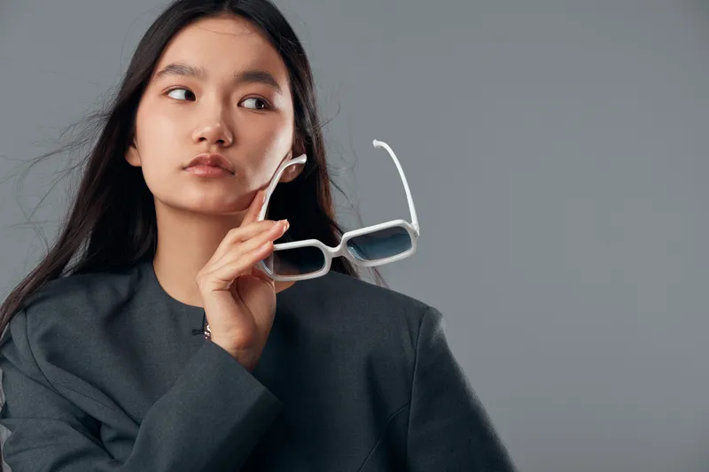 Woman with long black hair holding sunglasses in hand looking sideways, wearing dark blazer with serious expression on gray background in studio portrait photography.