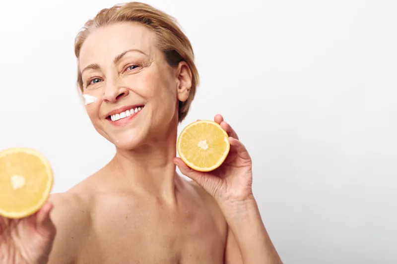 Smiling middle aged woman with natural skin holds two halves of fresh lemon, applying skincare cream strip on face. Concept of healthy lifestyle, vitamin care, and natural beauty wellness.