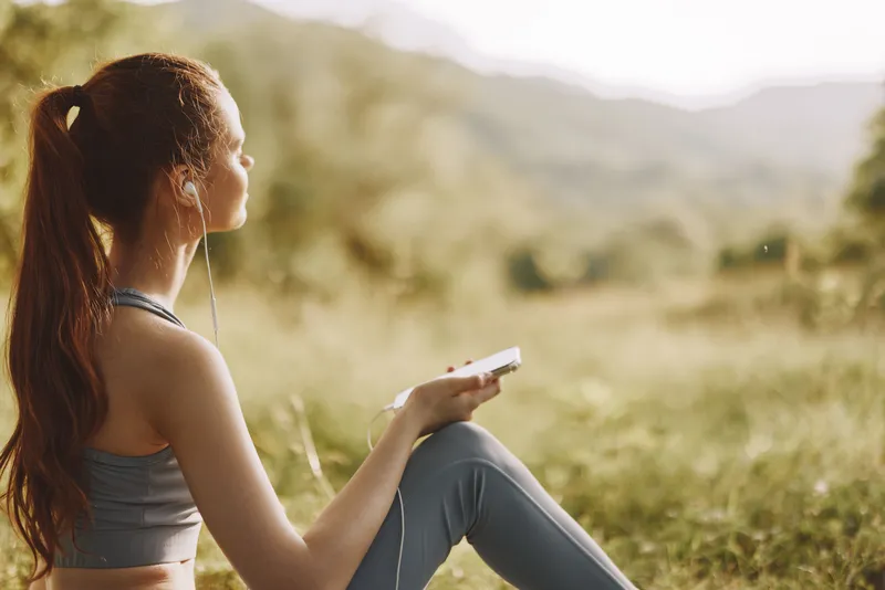 Serene young woman enjoying music during outdoor exercise in nature, wearing sports attire, with a soft focus on a beautiful green background