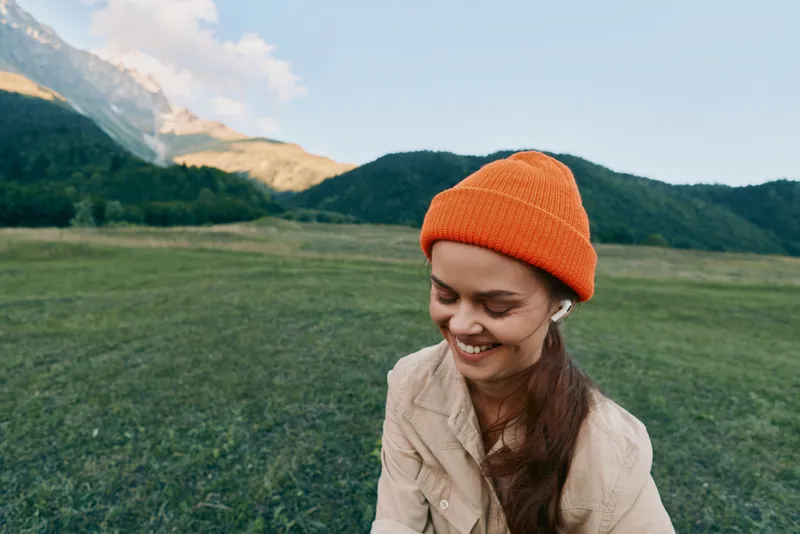 Young woman wearing an orange beanie and beige jacket smiles in a wide open field with green grass and distant mountains under a clear blue sky