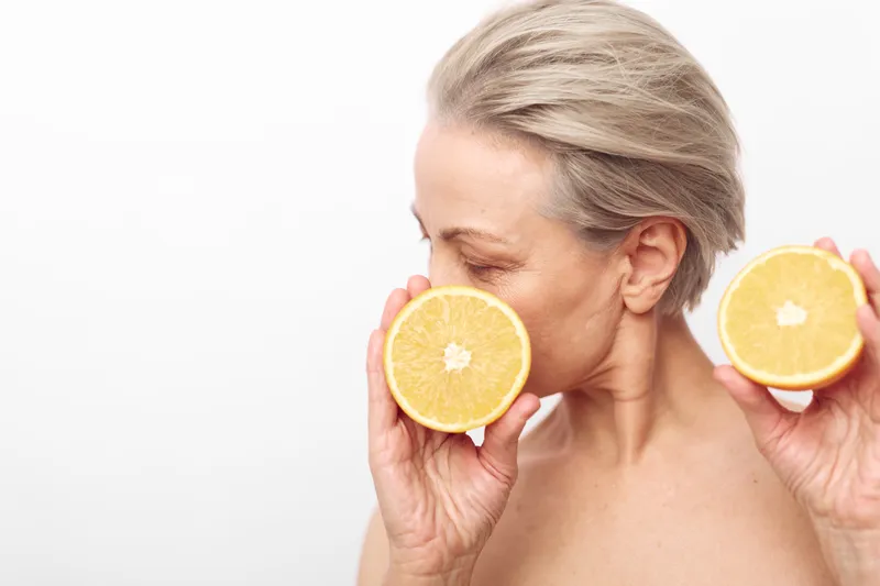 Mature woman with short grey hair holds two orange halves near her face against white background. Concept of natural beauty, healthy lifestyle, skincare and citrus fruits.