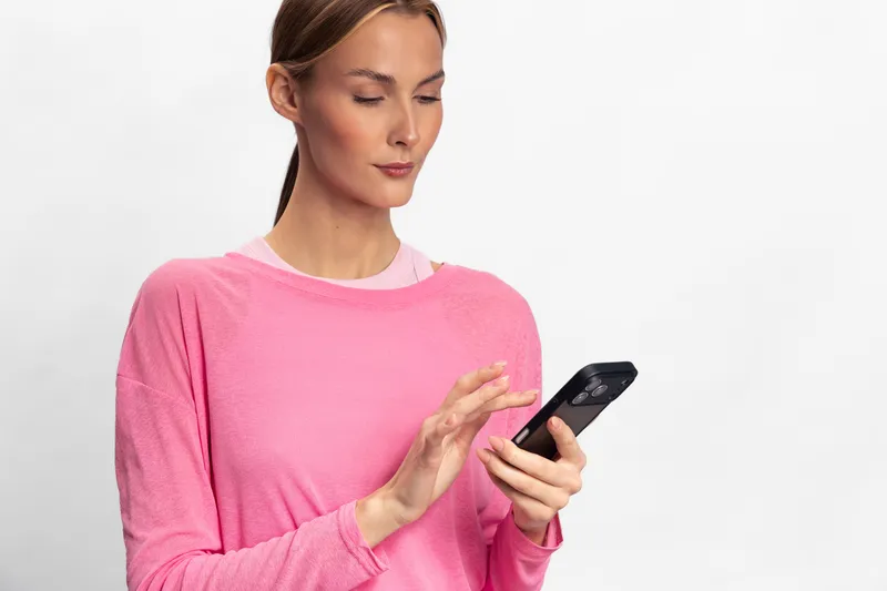 Young woman in pink sweater uses smartphone with focused expression on white background. Concept for technology, communication, and modern lifestyle with casual clothing.