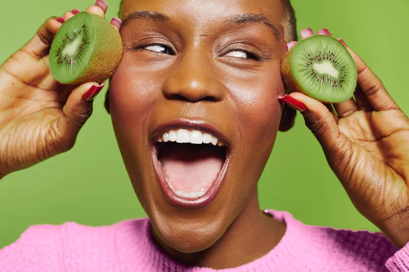 Happy African woman with dark skin holds two halves of kiwi fruit near face, smiling widely. Bright green background and pink sweater create fresh, joyful healthy lifestyle concept.