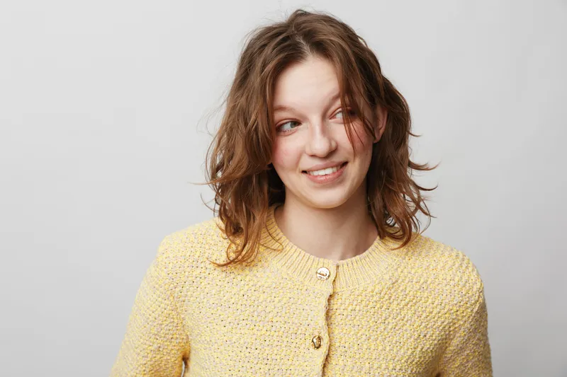 Young woman with wavy brown hair wearing a yellow knitted sweater smiles softly looking aside on a light grey background. Concept of casual style, happiness, and natural beauty.