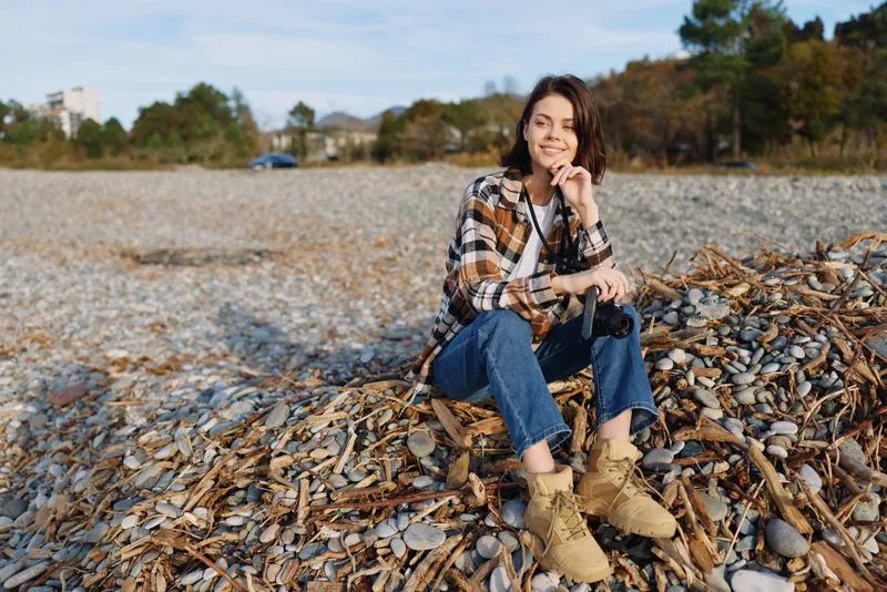 Woman sitting on a pebble beach in a plaid shirt and rugged boots, enjoying a sunny day by the shore with a calm coastal backdrop and distant trees behind.