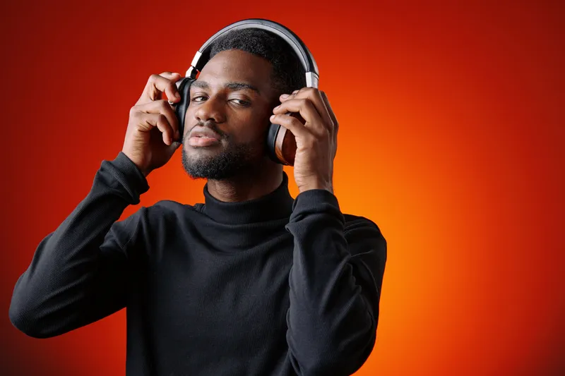 Young Black man enjoying music with headphones against a vibrant orange background, showcasing a relaxed and confident expression in modern attire