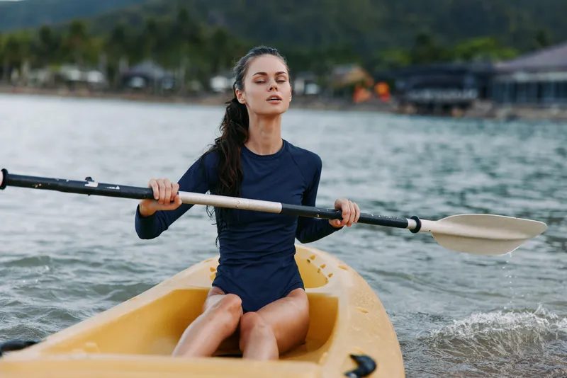 Young woman paddling a yellow kayak on calm water, gripping a double blade paddle with focus and balance as she glides toward the shoreline.