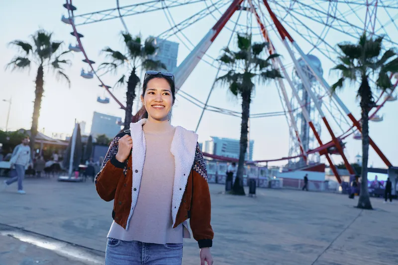 Woman at an outdoor amusement setting smiling beside a large ferris wheel, casual jacket and sweater, morning light, urban boardwalk vibe, carefree entertainment scene