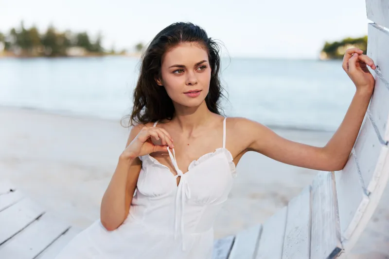A young woman stands beside a pale wooden railing on a sunny beach, wearing a white sundress, gazing softly into the distance with calm confidence.