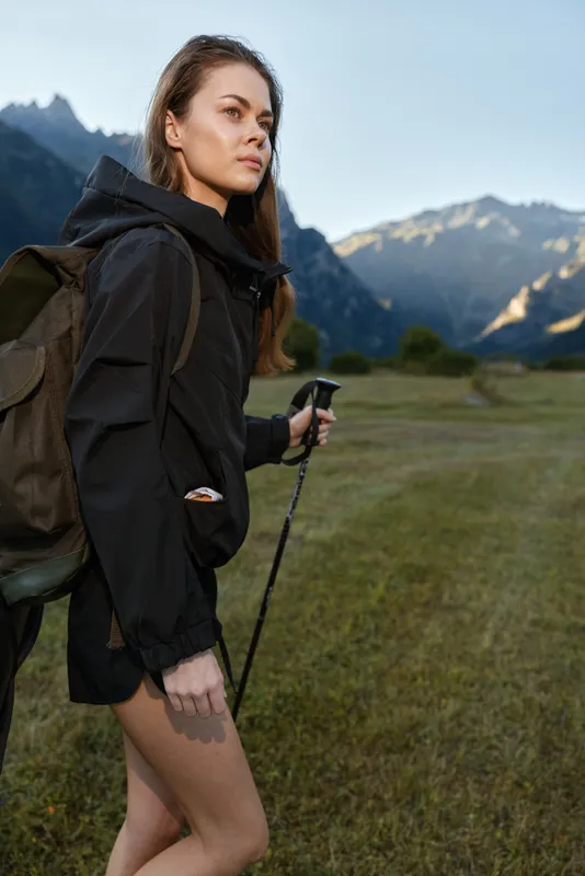 A young hiker with a backpack moves through a scenic meadow beside towering mountains. She holds a trekking pole and gazes forward with calm focus, enjoying fresh air, solitude, and a sense of