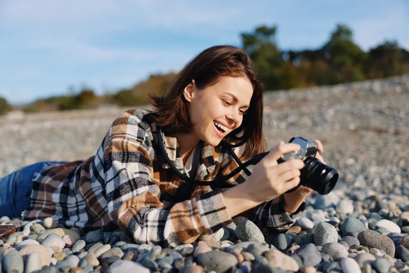 Woman lying on a pebble beach with a camera, smiling and focused, enjoying outdoor photography and leisure by the coast in casual plaid clothing, capturing a candid travel moment.