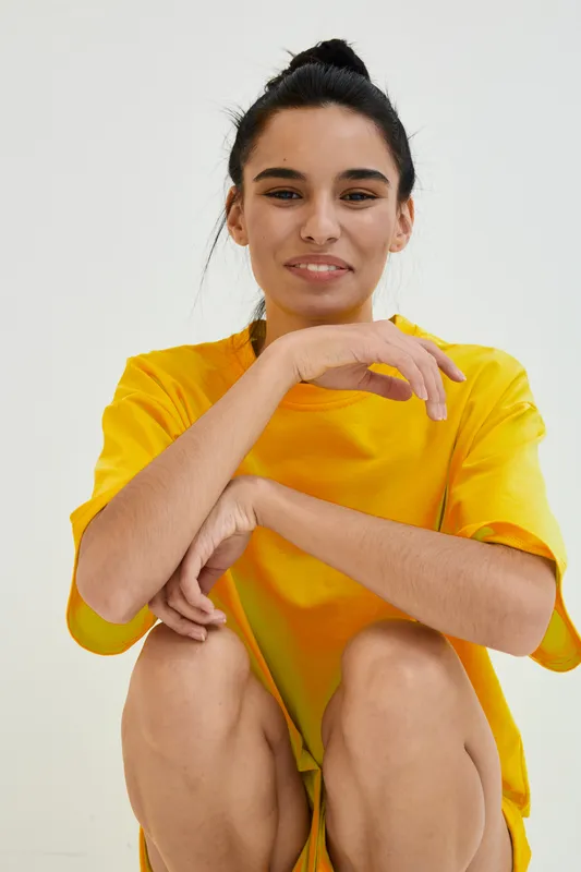 Young woman in yellow t-shirt smiling gently with casual hairstyle sitting against a light background, conveying warmth and relaxed mood in a simple minimalistic setting.