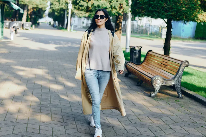 Fashion, woman, coat, street, park, walking, outdoors, style, casual, sunlight, sunglasses, elegant stroll in a beige coat paired with denim as she moves along a tree lined path in a peaceful urban
