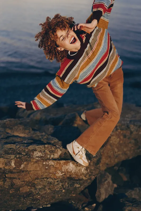 Woman lifestyle outdoors by the sea, standing on rocks, curly hair flying, smiling with happiness and laughter, relaxed pose in wind, sunlit sunset light, casual striped sweater, jeans boots
