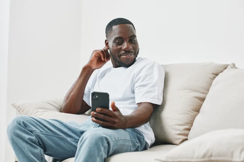 Young Black man enjoying music on his smartphone while relaxing on a light colored couch in a modern living room, conveying a sense of happiness and comfort
