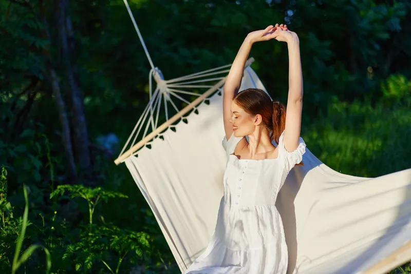 Young woman in a white dress relaxing in a hammock, enjoying nature on a sunny day, surrounded by greenery and warm light