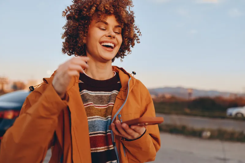 Woman lifestyle outdoors, laughing with curly hair in a casual jacket, sunlit golden hour, warm breeze, relaxed expression, near road and hills