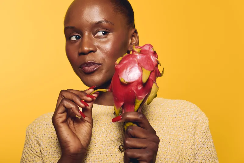 Young black woman with dark skin holds a red dragon fruit near her face, wearing a yellow sweater on a vibrant yellow background. Concept of healthy lifestyle and exotic fruit.