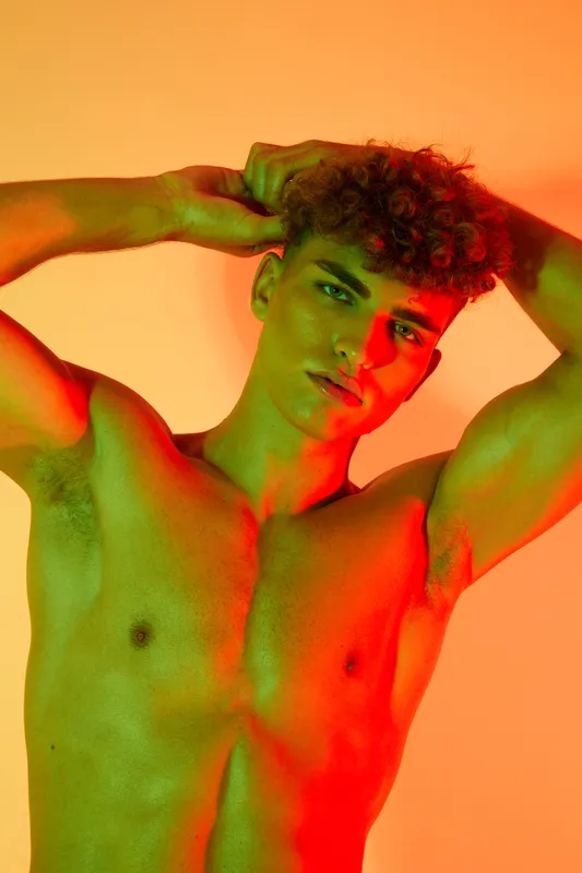 Young man with curly hair posing shirtless with hands behind head under colorful orange and green lighting for fashion, beauty and portrait photography concept.