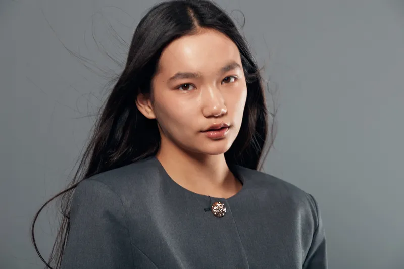 Portrait of young woman with long black hair wearing dark formal jacket with button looking at camera with neutral expression on gray background in studio lighting.