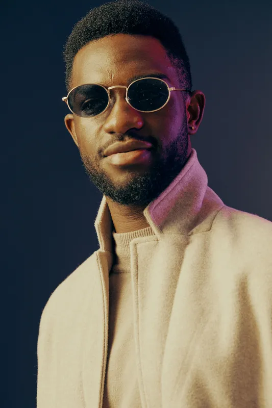 Stylish African American man in sunglasses and beige jacket, showcasing confidence and modern fashion against a dark blue background