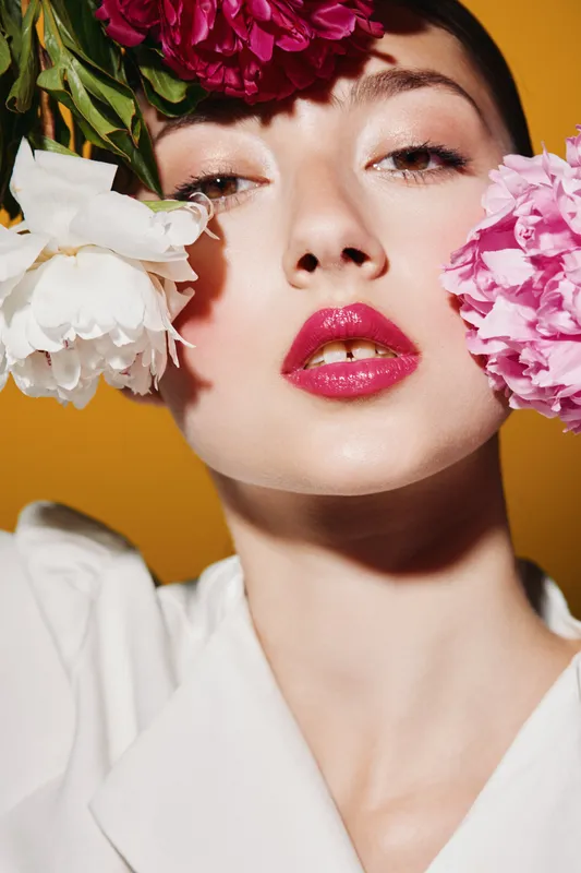 Close-up of a fashion woman with glossy lips holding peonies near her face on a yellow background. Creative beauty concept with floral accessories and luxury makeup.