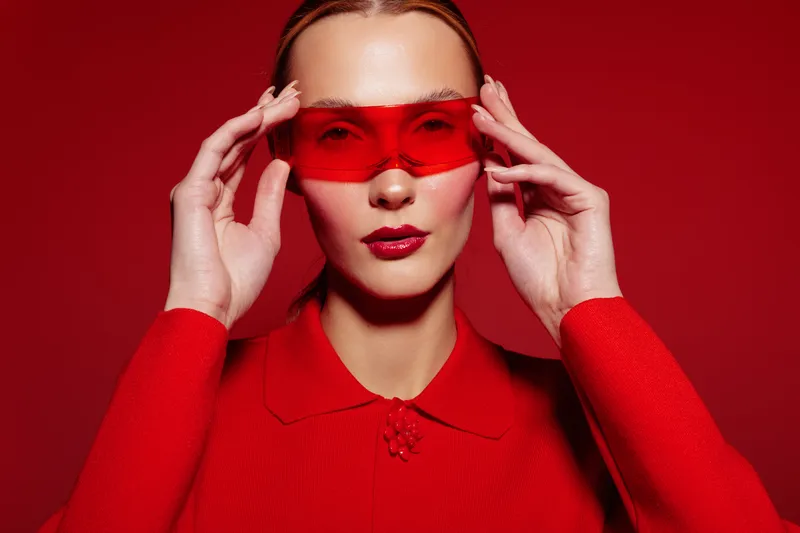 Stylish woman in red sweater and red sunglasses poses against red background. Fashion portrait with bold color contrast and confident expression. Concept of modern style and elegance.