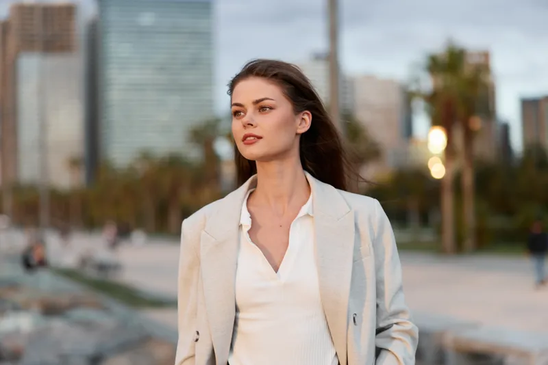 Confident woman in business attire walking in an urban park with palm trees and city skyline, showcasing a stylish and professional look, ideal for business themes.