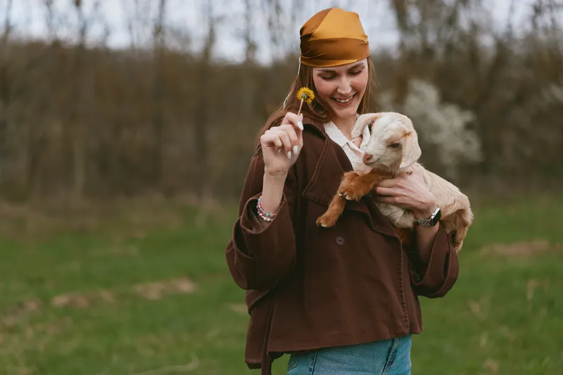 Cheerful woman outdoors, holding a goat kid, gentle smile, sunlit scene, casual attire, autumn tones, natural backdrop, playful moment, soft breeze, relaxed mood vibrant colors, open field, natural