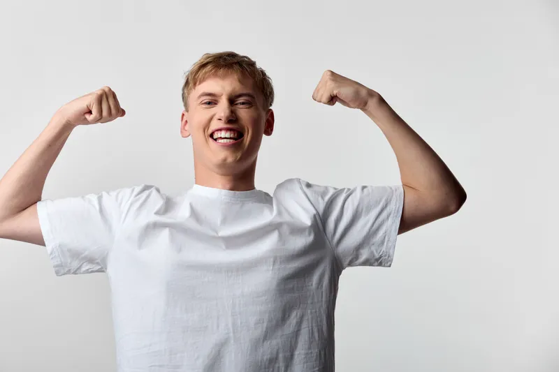 Flexing arms, confident smile from a man in a white tee, capturing energy and strength for fitness, wellness and motivation in a bright studio shot