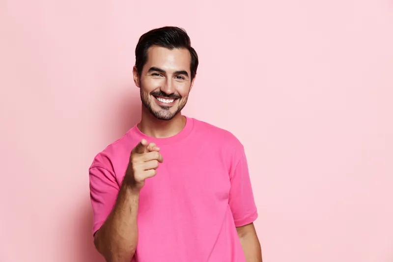 Smiling cheerful young man wearing a pink t-shirt pointing at the camera, isolated on a solid pastel pink background. Joyful mood and vibrant color expression.
