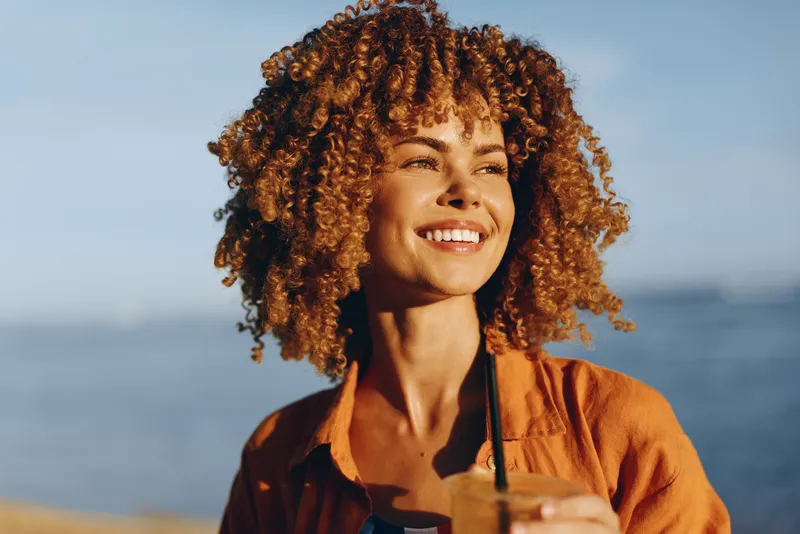 Smiling woman with curly hair seaside portrait, outdoors in warm sunlight, warm jacket, holding sunglasses, ocean horizon blurred background, cheerful head and shoulders closeup, natural lifestyle