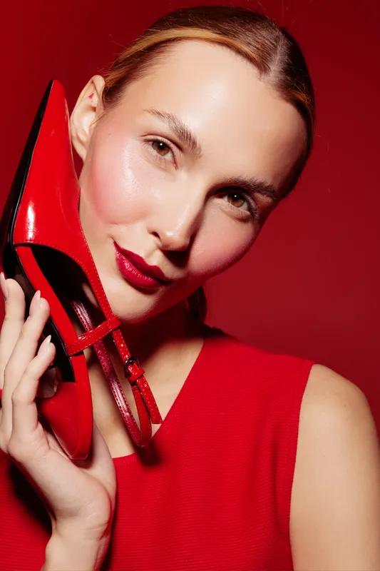 Close-up of a smiling woman in red dress holding a red high heel shoe near her face. Bright red background enhances the fashion concept and stylish beauty portrait.