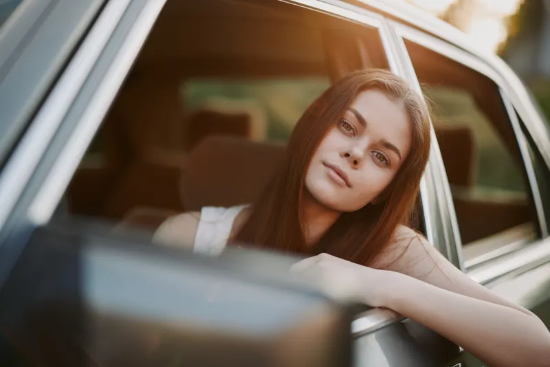 Young woman with long hair enjoying the sunset from a car window, embodying freedom and relaxation in a vibrant summer atmosphere.