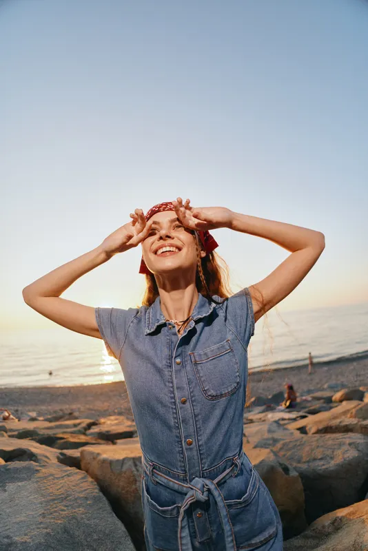 Happy woman in modern boho-western denim street style enjoys sunset on the beach with natural film color tones, expressing freedom and joyful lifestyle mood.