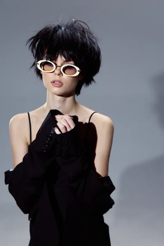 Stylish woman with black hair and white sunglasses wearing dark outfit posing against gray background