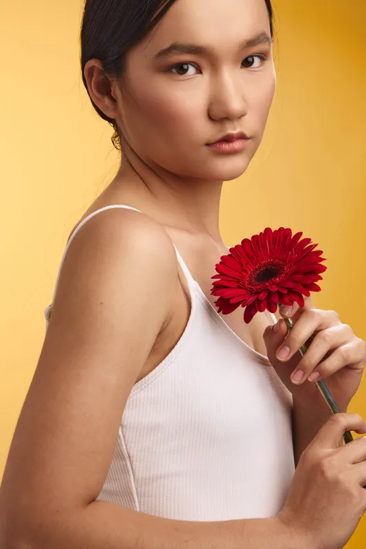 Asian woman is seen holding a red flower against a warm backdrop, exuding calm elegance and gentle confidence that invites a fresh, contemporary mood and graceful beauty.