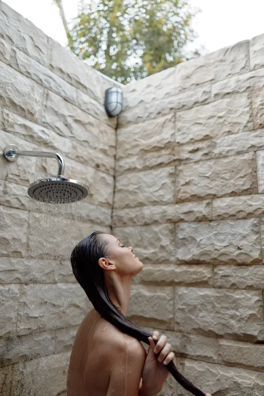 woman showering outdoors with long wet hair under rain showerhead in stone wall bathroom, natural lighting, relaxation, calm mood, wellness moment