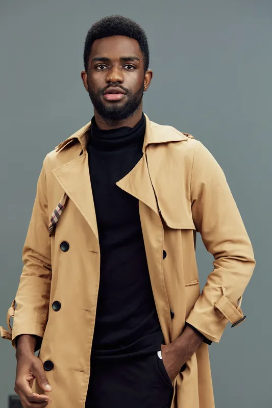 Fashionable black man in a stylish trench coat and black turtleneck on a muted background, exuding confidence and modern style