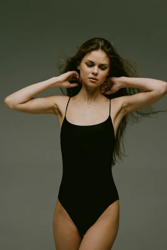 Woman in black swimsuit posing with hands near neck on neutral gray background