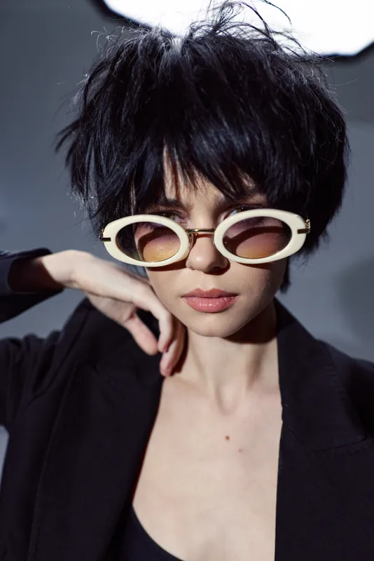 Fashionable young woman with short black hair wearing stylish sunglasses and a dark outfit in a minimalistic setting.