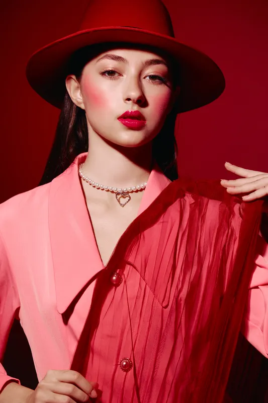 Stylish woman in a red hat and pink blazer poses with vibrant makeup and pearl necklace, showcasing bold fashion and creative beauty in a bright red studio scene.