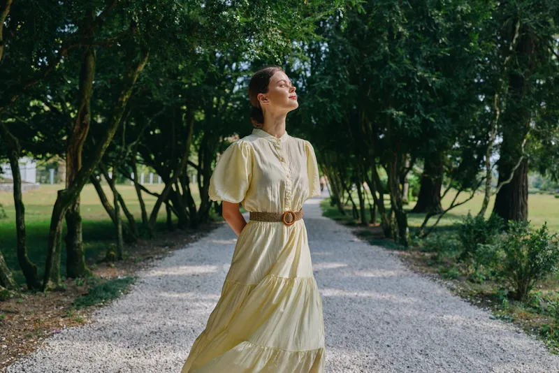 woman portrait in nature outdoors on tree lined path, long dress and sunlight, serene expression and authenticity inclusivity for lifestyle