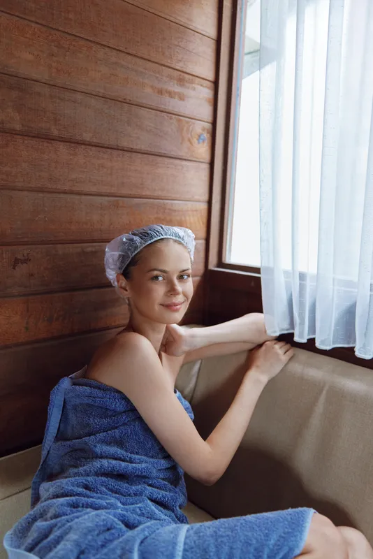 Young woman wrapped in blue towel and wearing shower cap sits by window in wooden spa room, showing calm and relaxed expression with soft natural light on skin.