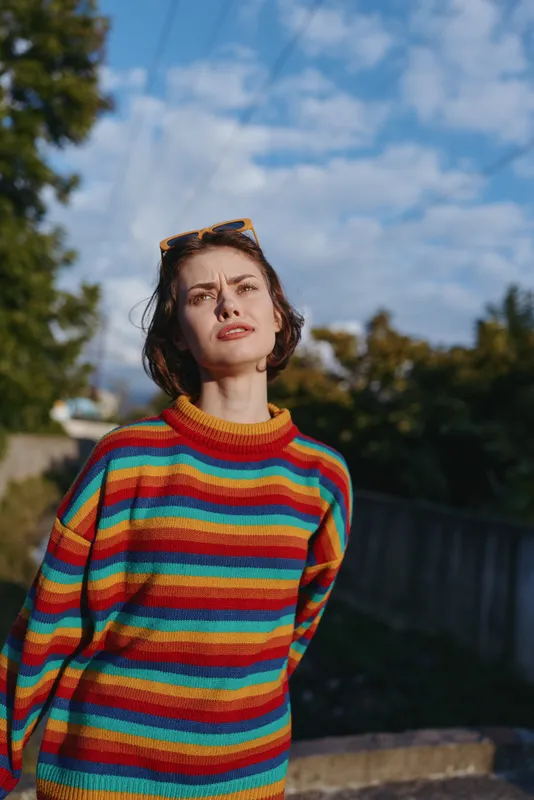 Woman in striped sweater, young adult with short hair and sunglasses, standing outdoor under blue sky in casual outfit, leaning slightly with thoughtful expression, colorful lifestyle portrait.