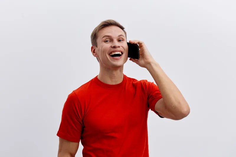Cheerful young man with short blond hair wearing red t-shirt talking on mobile phone smiling joyfully isolated on plain white background studio portrait. People lifestyle concept