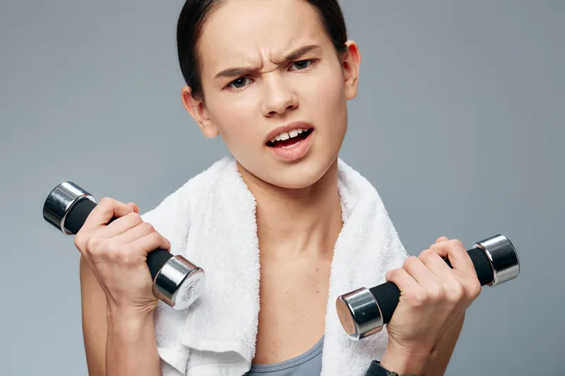 Female fitness enthusiast lifts dumbbells with towel after workout, focused expression, indoor gym backdrop, showcasing strength, effort and determination in a dynamic training session.