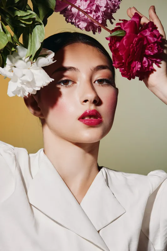 Woman with glossy lipstick and flawless skin surrounded by blooming peonies. Studio portrait with soft shadows and elegant fashion styling for beauty creative concept.