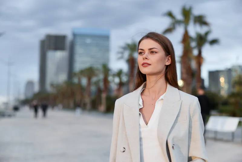 confident woman in beige coat walking outdoors with urban cityscape background during cloudy day, modern lifestyle and fashion concept