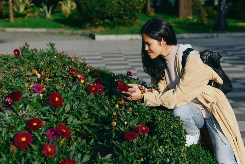 Woman kneeling beside vibrant red flowers in a garden, gently inspecting blossoms, enjoying nature, casual outdoor scene, lifestyle photography focused on gardening, spring colors, fresh air, serene